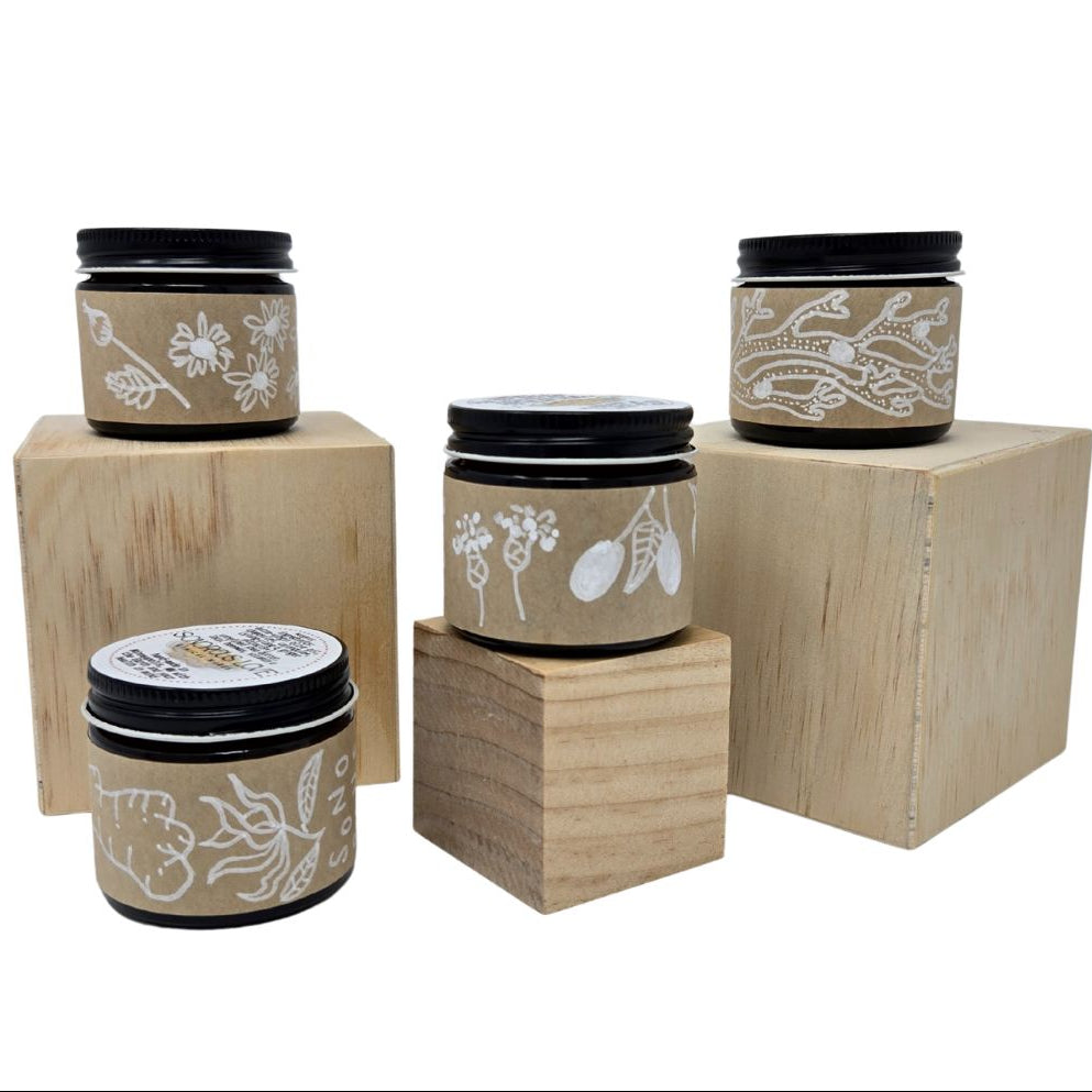 Set of four amber jars with herbal balms and hand-drawn product labels placed on wooden blocks against a white background