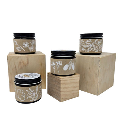 Set of four amber jars with herbal balms and hand-drawn product labels placed on wooden blocks against a white background