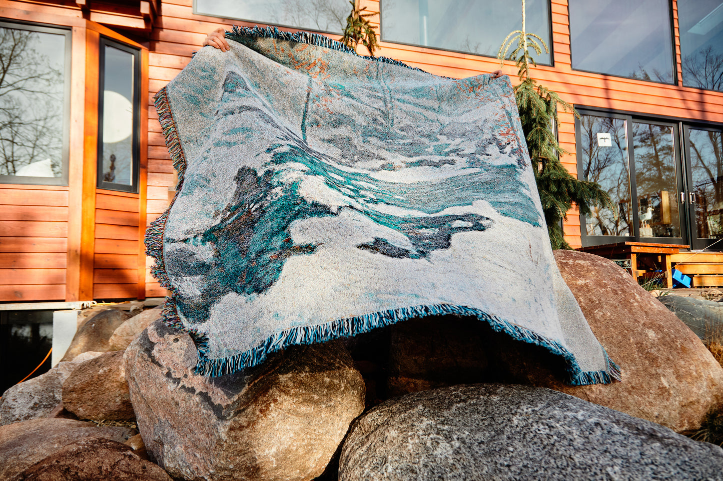 Icebound by John Henry Twachtman, featuring a serene abstract artwork nature scene with curves of ice and snow; white, blue and orange leaves. eco-friendly cotton woven blanket. Person holding up blanket over a white background. Person holding blanket up while standing on rocks in front of a home.