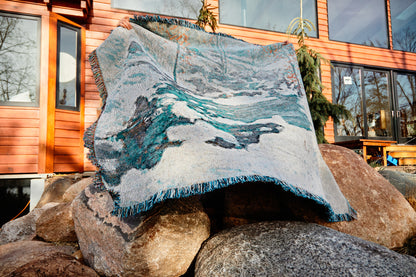 Icebound by John Henry Twachtman, featuring a serene abstract artwork nature scene with curves of ice and snow; white, blue and orange leaves. eco-friendly cotton woven blanket. Person holding up blanket over a white background. Person holding blanket up while standing on rocks in front of a home.